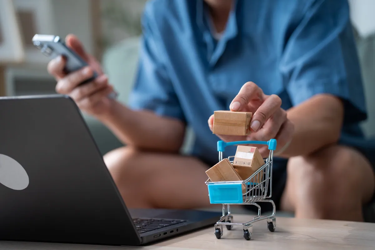 Une personne faisant des achats en ligne avec un téléphone, un ordinateur et un chariot miniature rempli de colis